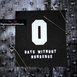 Könnte beinhalten: Schwarzer Stoff-Patch mit weißem Text, der "0 TAGE OHNE UNSINN" lautet. Die Zahl 0 ist in einer großen weißen Schrift. Der Patch ist auf einem schwarzen Hintergrund mit Sicherheitsnadeln befestigt.