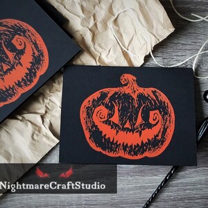 Puede incluir: Dos tarjetas negras con una ilustración roja y negra de una calabaza tallada. Las tarjetas están sobre un fondo de papel marrón.