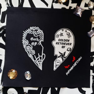 May include: Black fabric patch with a white embroidered heart design. The heart is split in half, with one side saying "Goth GF" and the other side saying "Golden Retriever BF".