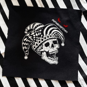 May include: Black square patch featuring a white skull wearing a jester's hat with a black and white checkered pattern. The patch has the text "nightmarecraftstudio" in white with two red bat silhouettes.