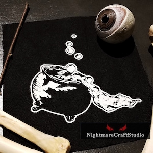 May include: Black and white embroidered patch of a cauldron with bubbling liquid and steam. The patch is on a black fabric background. The text "NightmareCraftStudio" is at the bottom of the patch.
