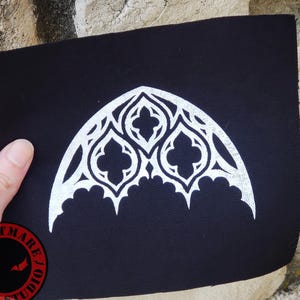 May include: Black fabric patch featuring a white gothic arch design. The patch has a decorative, symmetrical pattern with pointed arches and quatrefoils. The patch is held up by a hand, with a red and black logo in the corner.
