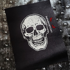May include: A black patch featuring a detailed white skull design. The skull has visible teeth and eye sockets. The text "NIGHTMARECRAFTSTUDIO" is printed on the right side of the patch in white.