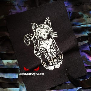 Könnte beinhalten: Schwarzer Stoffaufnäher mit einem weißen, stilisierten Katzendesign und einem Mond. Die Katze sitzt mit eingerolltem Schwanz. Der Text "NIGHTMARECRAFTSTUDIO" ist unter der Katzenillustration in Weiß gedruckt.