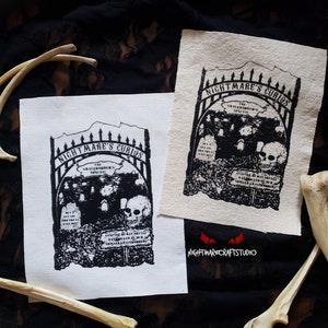 Op de afbeelding: Twee witte stoffen patches met zwarte inkt geprinte ontwerpen. Het ontwerp is een poort met de woorden "Nightmare's Curios" erboven. De poort staat open en er zijn schedels en grafstenen in te zien. De tekst "The Graveyard's Spirits" is in de poort gedrukt. De tekst "Genuine Whispers May Smell Like Rotting Flesh" is op een bord onder de poort gedrukt. De tekst "Nightmare Crafts Studio" is onder de patches gedrukt.