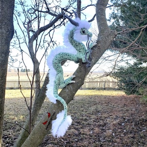 May include: A handmade, green and white crochet dragon with a long, flowing body and a fluffy white mane and tail. The dragon is perched on a tree branch, with a detailed face and horns. The dragon is a unique, whimsical decorative item.