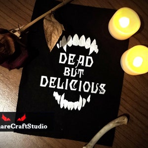 Puede incluir: Parche de tela negro con texto blanco que dice "Dead But Delicious" y una imagen blanca de un conjunto de colmillos. El parche está sobre una superficie de madera con una rosa seca y dos velas encendidas.