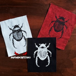 May include: Three fabric patches featuring a black and white illustration of a beetle on each. The patches are displayed on a wooden surface. The patches are on white, red, and black fabric.
