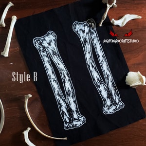 May include: Black fabric patch with two white skeletal arm bones printed on it. The text "Style B" is printed in white on the fabric. The patch is surrounded by white bones on a wooden surface.