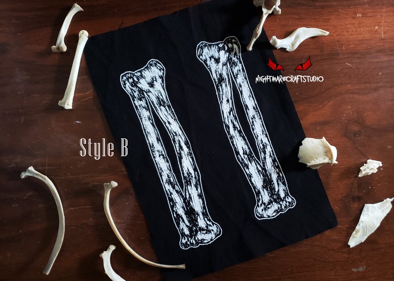 May include: Black fabric patch with two white skeletal arm bones printed on it. The text "Style B" is printed in white on the fabric. The patch is surrounded by white bones on a wooden surface.