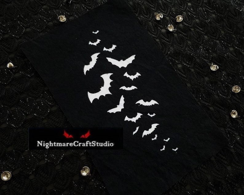 May include: Black fabric patch with a white embroidered design of bats flying in a diagonal line. The patch has the text "NightmareCraftStudio" in red letters.