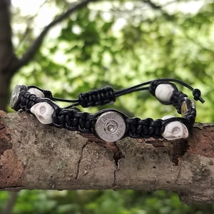 May include: A black paracord bracelet with white skull beads and a silver bullet charm. The bracelet is on a tree branch.