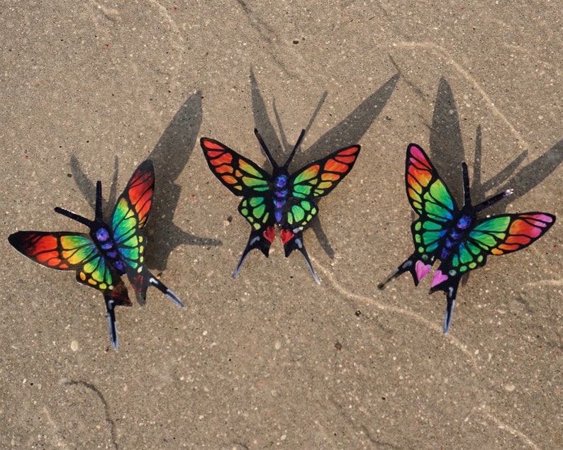 May include: Three colorful butterflies with iridescent wings. The butterflies are arranged in a triangular pattern on a sandy surface. The butterflies are red, green, blue, and yellow.