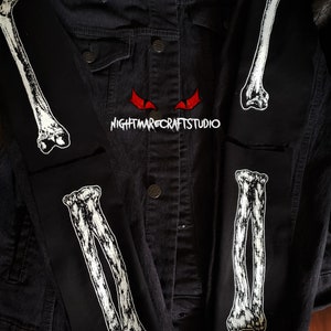 May include: A black denim jacket with white bone patches on the sleeves. The patches are of a human arm bone. The text "NIGHTMARECRAFTSTUDIO" is printed on the jacket.
