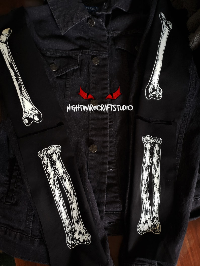 May include: A black denim jacket with white bone patches on the sleeves. The patches are of a human arm bone. The text "NIGHTMARECRAFTSTUDIO" is printed on the jacket.