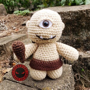 Puede incluir: Muñeco cíclope de ganchillo hecho a mano en beige y marrón. El muñeco tiene un ojo grande, una sonrisa con dientes y un garrote marrón. Lleva un cinturón marrón. Se ve un logotipo rojo y negro.