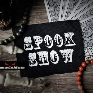 Könnte beinhalten: Schwarzer Stoff-Patch mit weißer Aufschrift "SPOOK SHOW". Der Patch ist von verschiedenen Objekten umgeben, darunter ein Schädel, Perlen und Spielkarten.