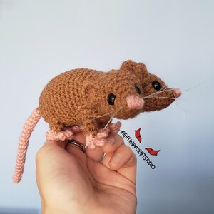 May include: Crocheted brown mouse with a pink tail, ears, and feet. The mouse has black eyes and long white whiskers. The mouse is handmade and appears to be a toy or decorative item.