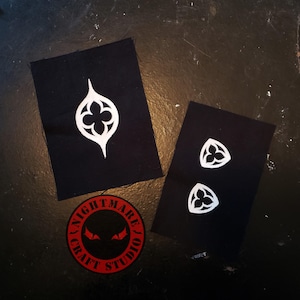 May include: Black fabric patches with white gothic-style designs. One patch features a large, stylized floral shape, while another has two smaller versions. A red circular patch with the text "NIGHTMARE CRAFT STUDIO" and stylized eyes is also present.