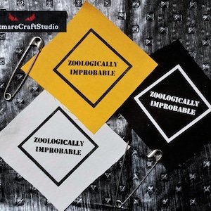 Pode incluir: Três patches para passar a ferro com forma de diamante e contornos pretos. Cada patch tem o texto "Zoologicamente improvável" em letras brancas. Os patches estão sobre uma superfície preta texturizada com alfinetes de segurança.