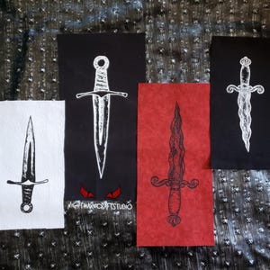 以下が含まれることがあります： 黒、赤、白の背景に白い刺繍で描かれた短剣の画像が施された、異なる4つの布製パッチ。パッチはすべて長方形で、少し使い古したような外観をしています。1つのパッチに「NIGHTMARECRAFTSSTUDIO」というテキストが表示されています。