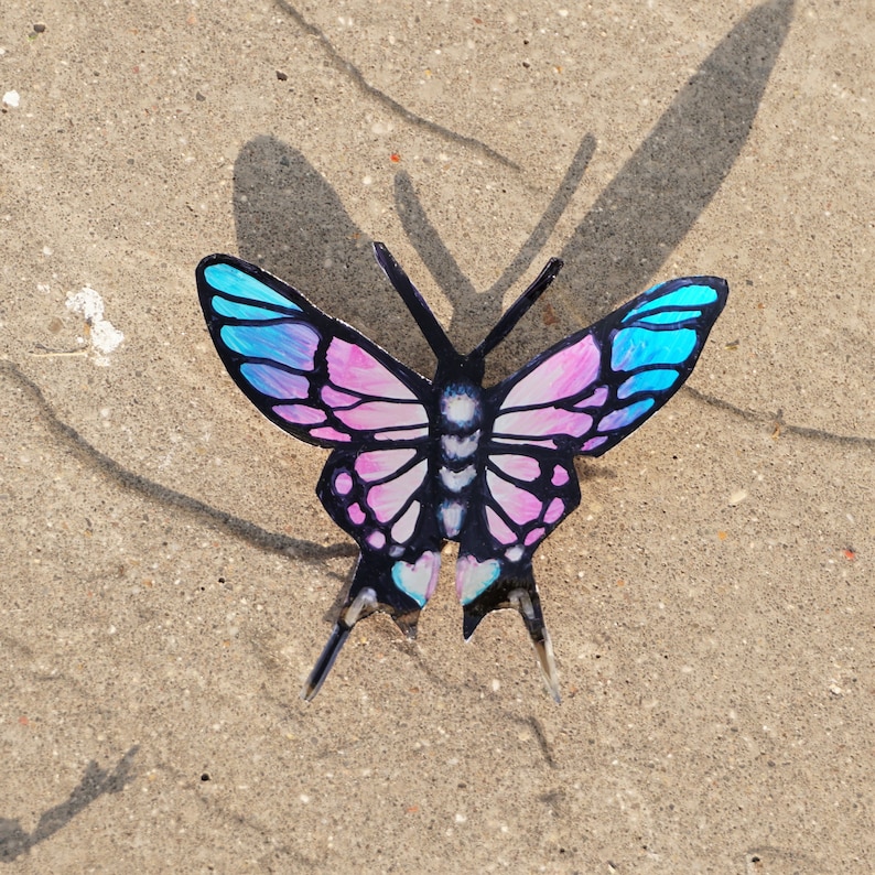 May include: A pink and blue butterfly with black markings on a sandy background.
