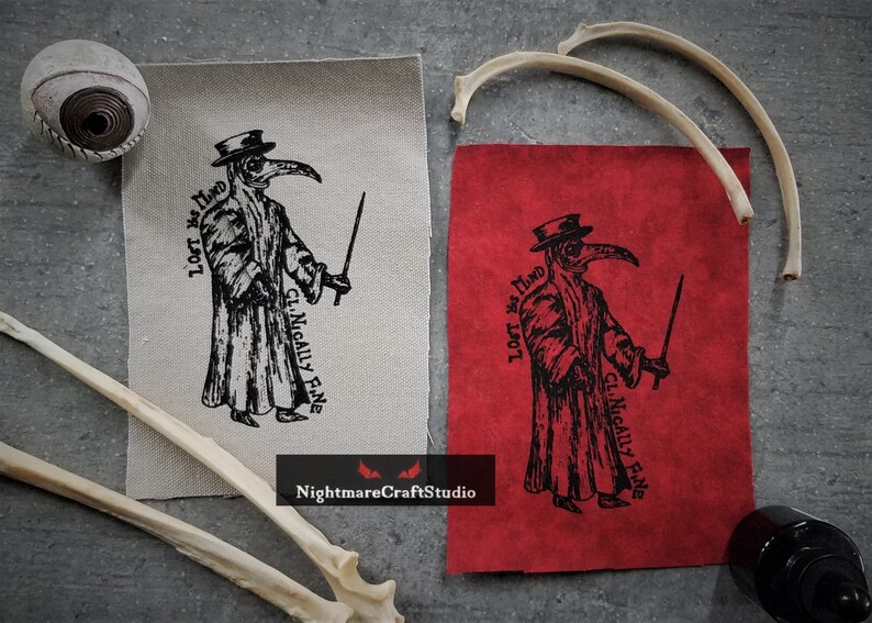 May include: Two fabric patches with a black and white illustration of a plague doctor holding a staff. The text on the patches reads "Lest We  Find" and "Clinically Fine".