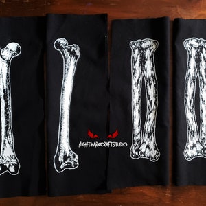 May include: Four black fabric patches with white printed images of human bones. The patches are sewn together and feature the text "NIGHTMARECRAFTSTUDIO" in red.