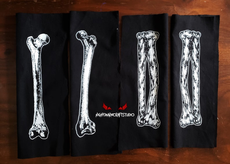 May include: Four black fabric patches with white printed images of human bones. The patches are sewn together and feature the text "NIGHTMARECRAFTSTUDIO" in red.