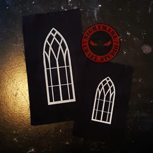 May include: Two white gothic window cutouts on black fabric. The larger window is approximately 6 inches tall, and the smaller one is about 3 inches tall. A red and black circular logo with the text "NIGHTMARE CRAFT STUDIO" is also visible.