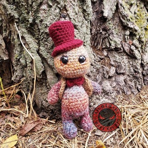 Może przedstawiać: Ręcznie robiona szydełkowa lalka z bordowym cylindrem i muszką. Lalka ma duże czarne oczy i jest wykonana z melanżowej włóczki w odcieniach brązu, czerwieni i niebieskiego. Stoi przed drzewem.