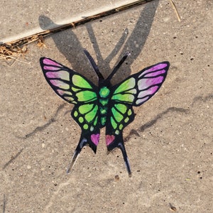 May include: A green and purple butterfly with black veins and pink hearts on its wings. The butterfly is on a gray concrete surface.