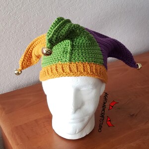 May include: A green, purple, and yellow crocheted jester hat with gold bells. The hat is on a mannequin head.