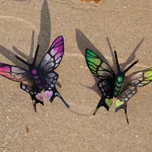 May include: Two decorative metal butterflies with black wings and painted details. One butterfly has pink and white wings, the other has green and white wings.