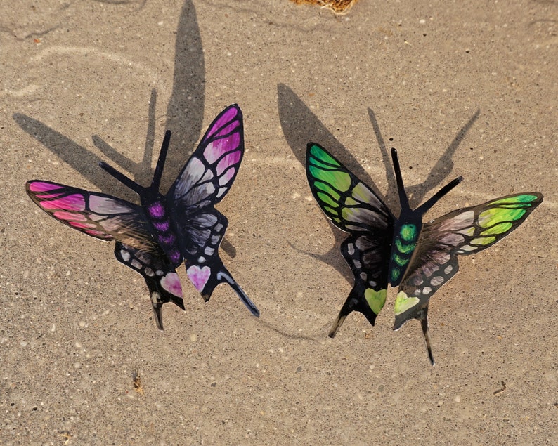 May include: Two decorative metal butterflies with black wings and painted details. One butterfly has pink and white wings, the other has green and white wings.