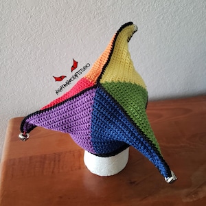 May include: A multi-colored crochet jester hat featuring four distinct points in yellow, green, blue, and purple, each tipped with a small silver bell. The hat is outlined in black yarn and includes the text NIGHTMARECRAFTSTUDIO with red eye icons.