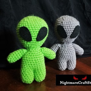 Peut inclure: Deux jouets en peluche extraterrestres en crochet. L'un est vert et l'autre est gris. Les deux ont des yeux noirs et sont debout sur une surface en bois.