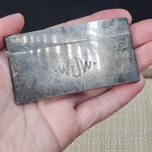 May include: A rectangular silver-toned metal case with a hinged lid and a weathered, scratched surface. The front of the case features an engraved monogram with the letters WJW centered within a diamond-shaped outline. The metal has a dull finish.