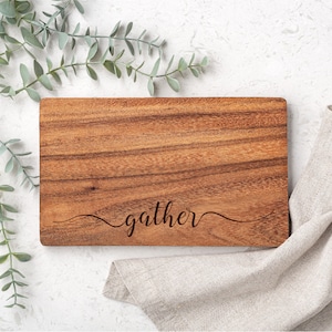 Puede incluir: Una tabla de cortar rectangular de madera con la palabra "gather" grabada en cursiva. La tabla es marrón con un patrón de vetas de madera natural. Una servilleta de lino beige y ramitas verdes con hojas están dispuestas alrededor de la tabla.