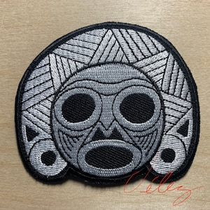 May include: Embroidered patch featuring a stylized tribal mask design in shades of gray and black. The patch has a black border and intricate geometric patterns, suitable for jackets, bags, or other fabric items.