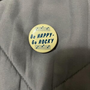 May include: A round, cream-colored button with the text "Be HAPPY-Go ROCKY" in blue, along with musical notes. The button is set against a gray, quilted fabric background.