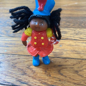 May include: A black Cabbage Patch Kid doll wearing a red and gold uniform with a blue hat and a candy cane. The doll has black yarn hair.