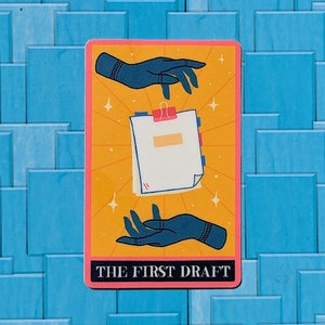 May include: A tarot card with a yellow background and a pink border. Two blue hands hold a stack of white papers with a yellow paperclip. The text "THE FIRST DRAFT" is printed on the card.