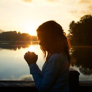Könnte beinhalten: Silhouette einer Person, die mit gefalteten Händen betet und auf einem Holzsteg steht, der einen See bei Sonnenuntergang überblickt.