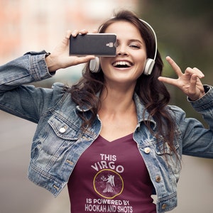 May include: A woman wearing a maroon V-neck t-shirt with the text "This Virgo is powered by hookah and shots" and a gold astrological symbol. She is wearing a denim jacket and headphones.