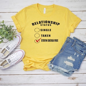 Op de afbeelding: Geel t-shirt met de tekst "RELATIONSHIP STATUS" en de opties "SINGLE", "TAKEN" en "STAYIN SUCKA FREE" met een vinkje. Het shirt wordt gecombineerd met een spijkerbroekje en witte sneakers op een houten ondergrond.