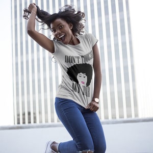 Puede incluir: Una mujer con una camiseta gris con el texto "Don't Touch My Hair" y una ilustración en blanco y negro de una mujer con un afro. Lleva pantalones vaqueros azules y está saltando en el aire.
