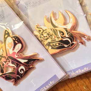 May include: Five enamel pins with black and white designs on a rose gold background. The pins feature different characters with wings and are labelled "dreamer", "crescent", "mourner", "trickster", and "mystic".