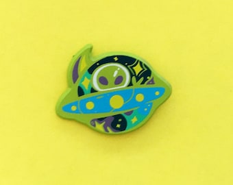 May include: Green enamel pin of an alien in a spaceship with a blue and yellow design. The spaceship has stars and swirls around it.