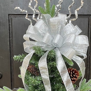 May include: A small artificial Christmas tree decorated with a silver ribbon bow and silver tinsel. The tree is decorated with pine cones.